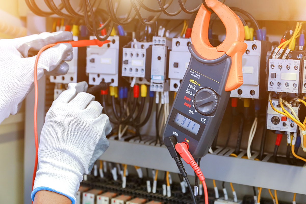 Electrician using a clamp meter to test voltage and current inside an electrical control panel.