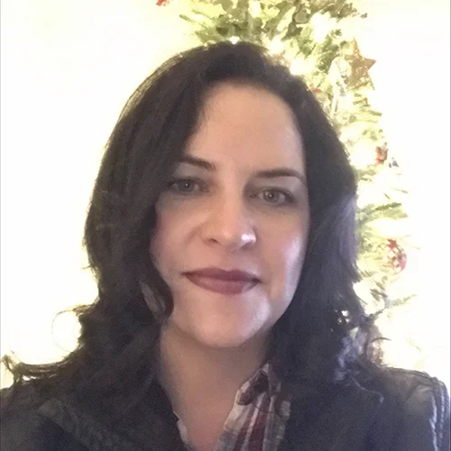 Woman with dark hair and red lipstick smiling, wearing a plaid shirt and leather jacket, with a Christmas tree in the softly lit background